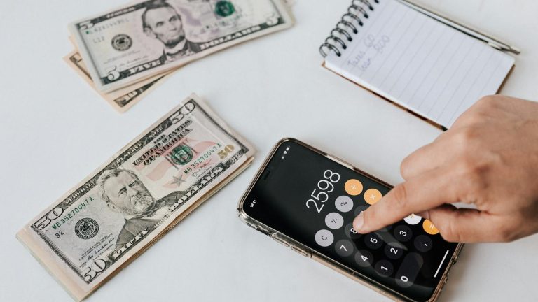 From above of crop anonymous economist calculating total amount of income using calculator app on cellphone near piles of greenbacks and notebook with pen